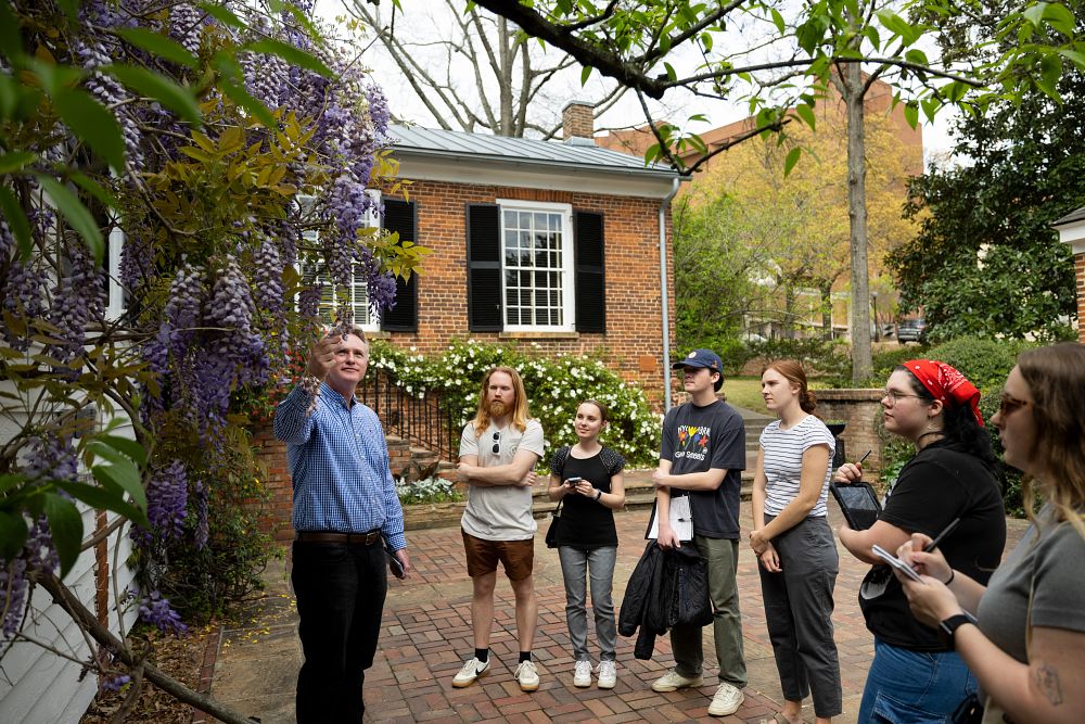 A professor holds a flower while teaching a group of students outside a building.